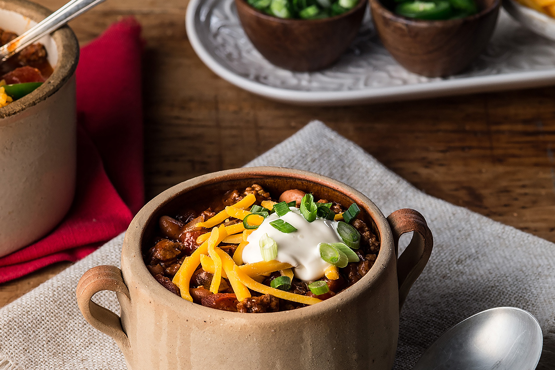 Bean and beef chili in small tan crock, garnished with shredded cheese, sour cream and green onions 