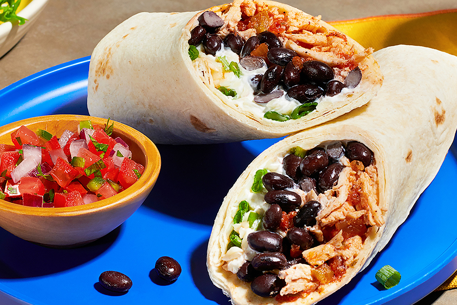 A chicken and black bean burrito sliced in half on a blue plate with small bowl of tomato salsa