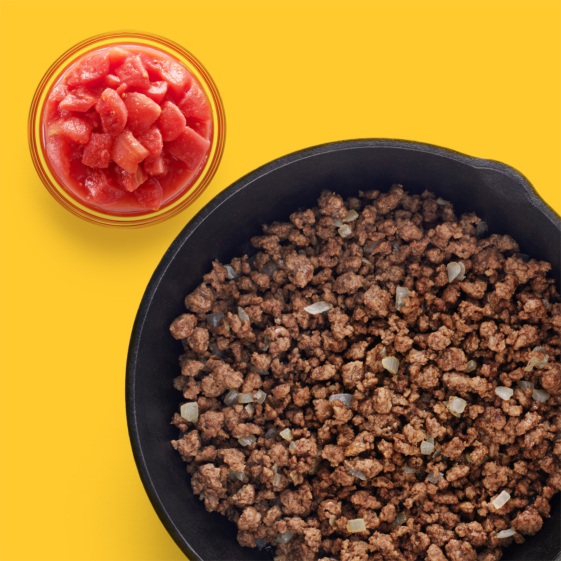 Cast iron pan full of browned ground beef with a yellow background and a side of tomatoes.