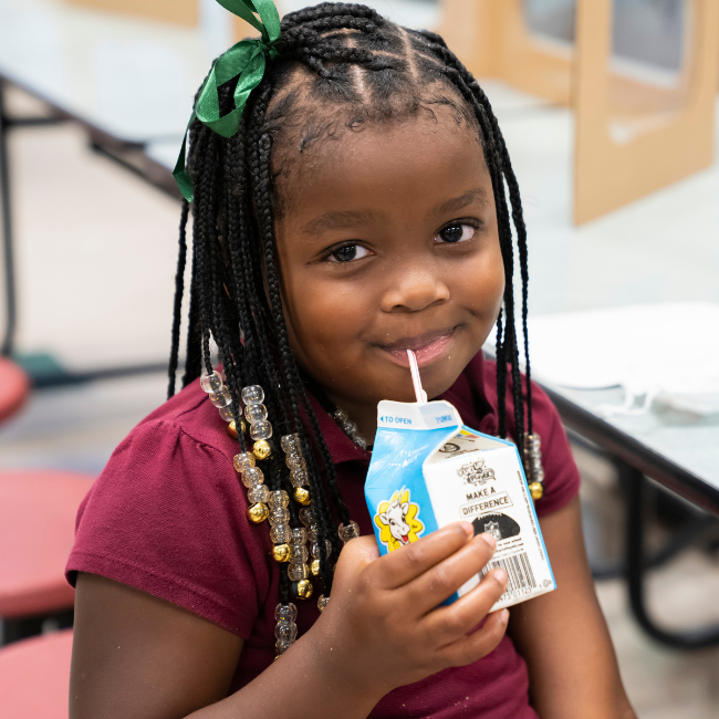 Elementary school kid drinking milk from a carton at a lunch table.