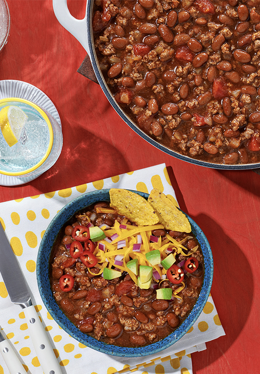 Bowl of chili next to a pot of chili on a red tablecloth