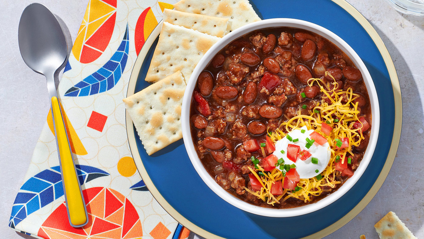 Chili in bowl with sour cream, cheese, and tomatoes, on a blue plate with saltines