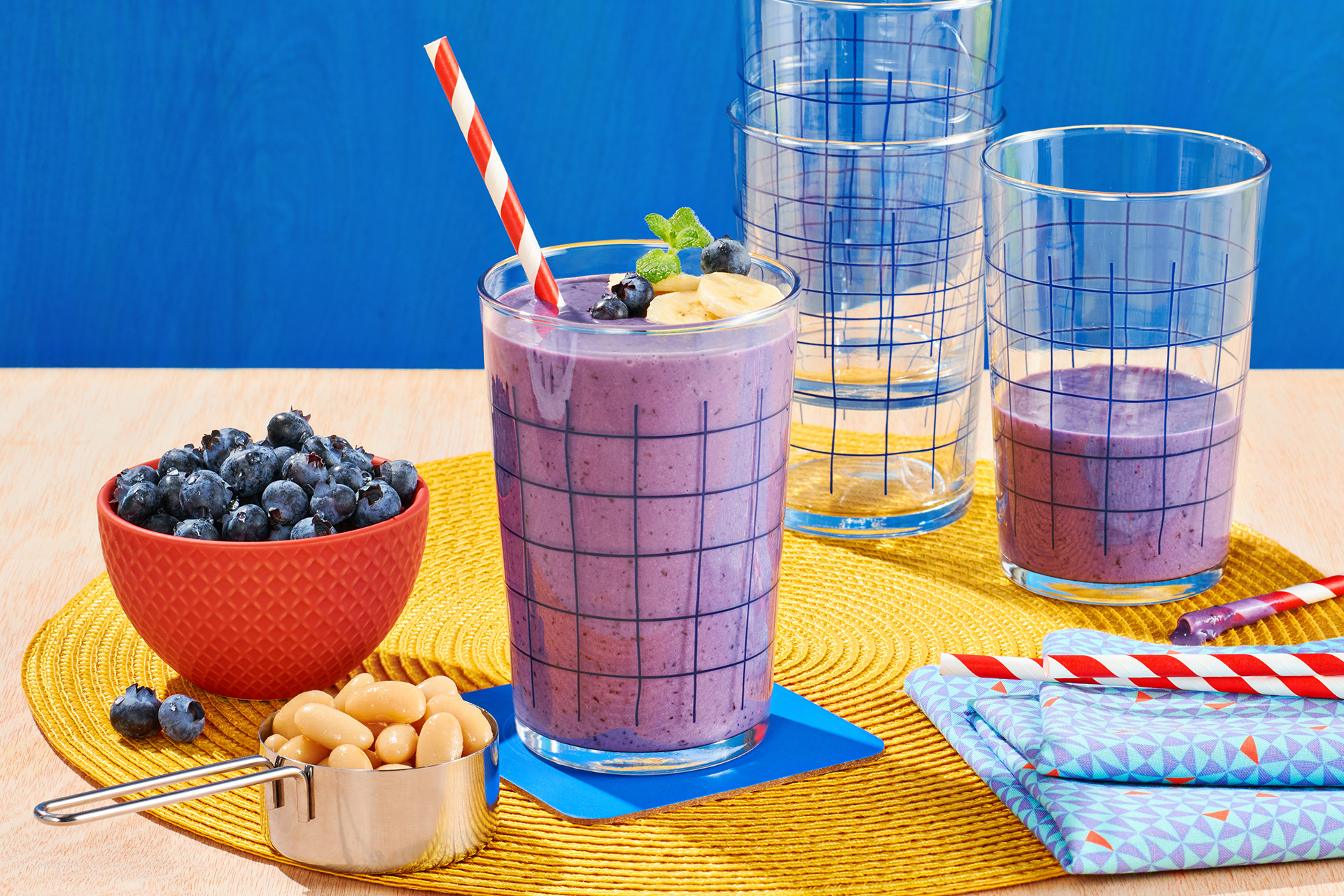Banana blueberry and bean smoothie with striped straw next to a red bowl filled with blueberries and a silver measuring cup filled with white beans
