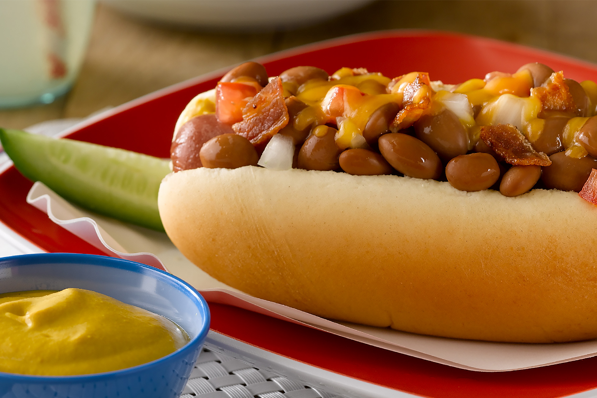 Close up of a cheddar chili dog with beans and bacon, with small bowl of mustard and pickle spear