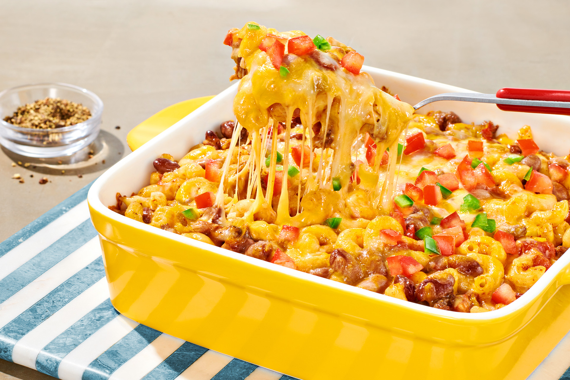 A spoon taking a scoop of a baked Mexican chili bean, macaroni and cheese casserole topped with tomato in yellow baking dish