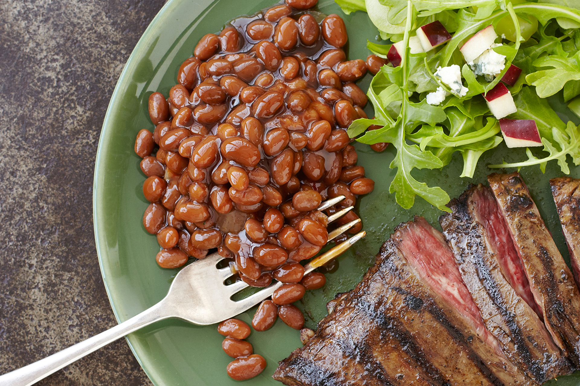 Sliced marinated steak with baked beans and a green salad with pear and bleu cheese on a white plate