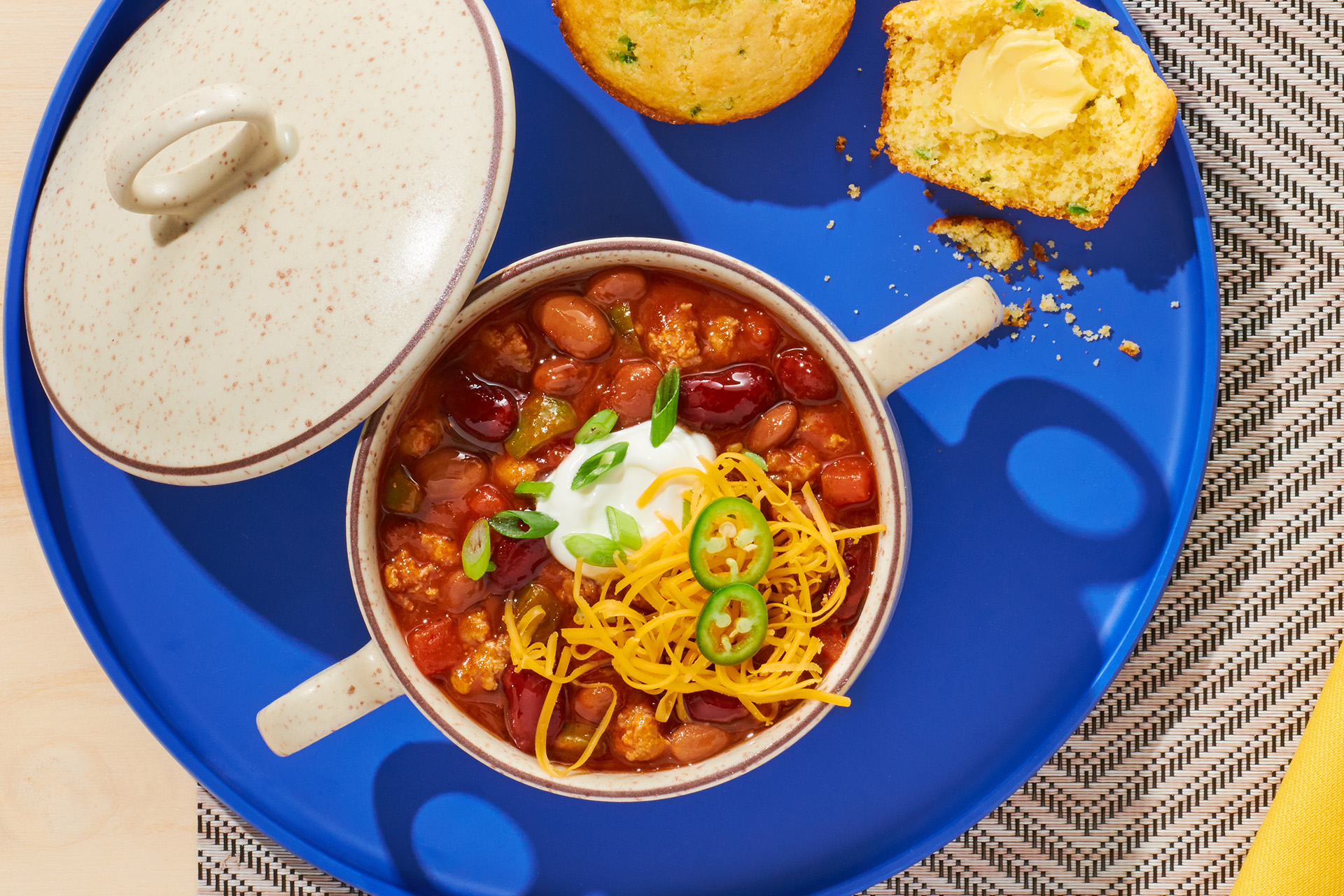 A bowl of chili topped with sour cream, jalapenos, and shredded cheese on a blue tray with corn muffins
