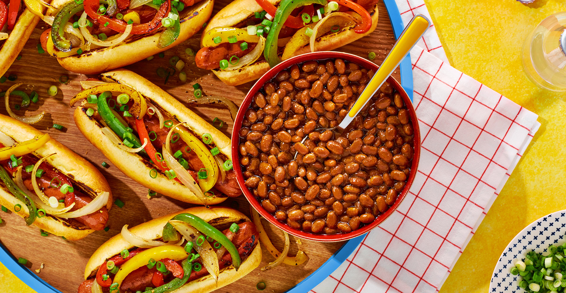 A blue serving tray loaded with several fajita dogs and a red bowl full of sweet head beans sitting on a yellow table.
