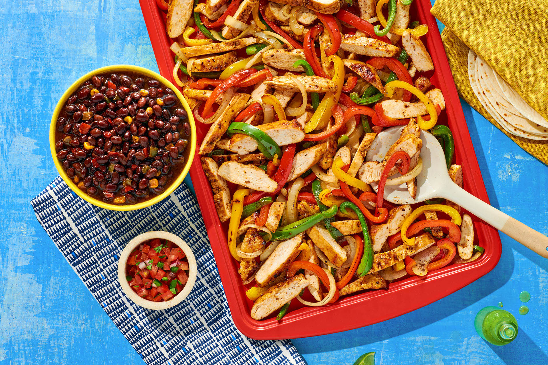 Fajita chicken and peppers on a red baking sheet next to a small bowl of beans on a blue table. 