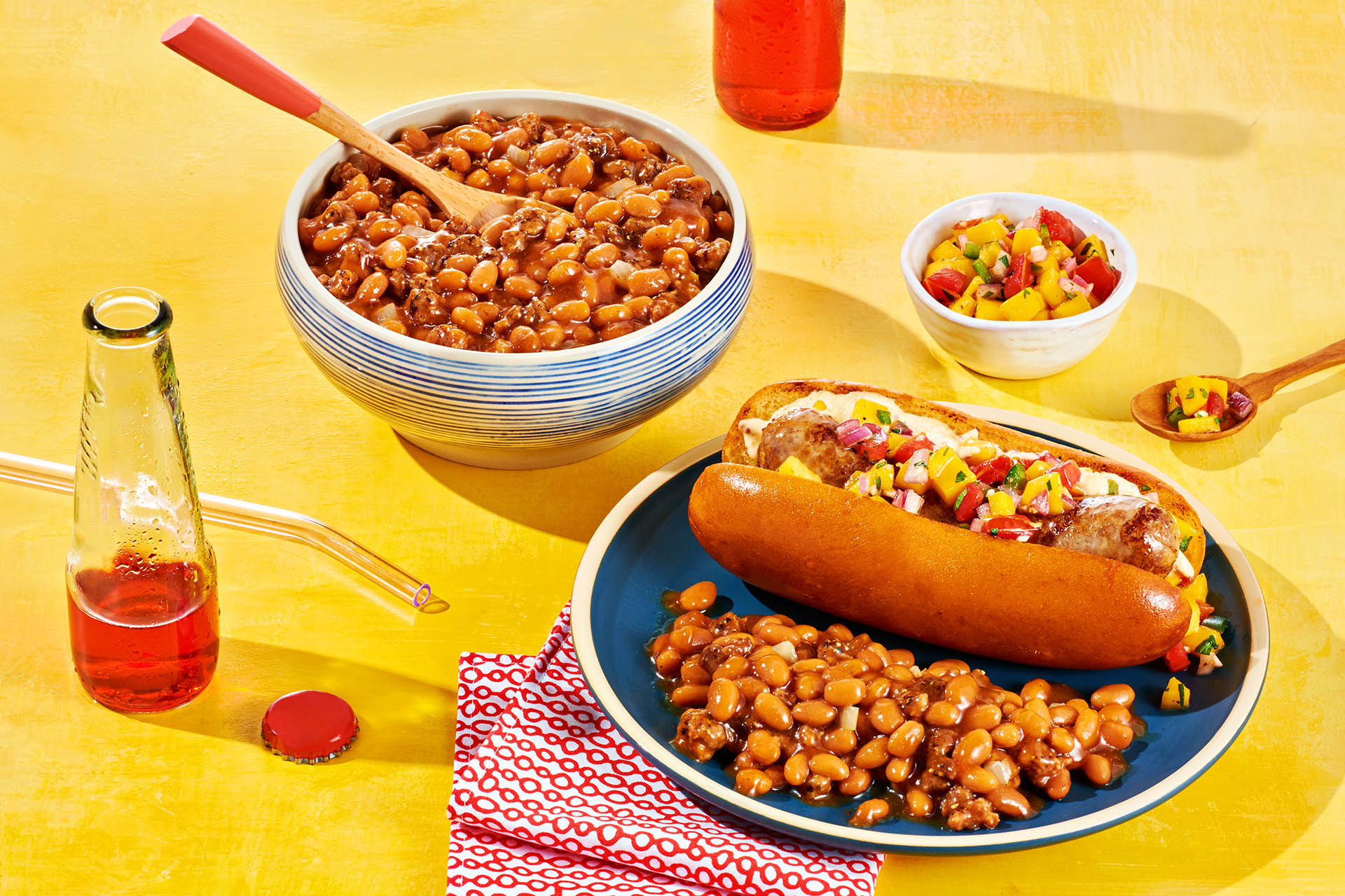A grilled brat on a bun with a side of hamburger baked beans next to bowl of hamburger baked beans.