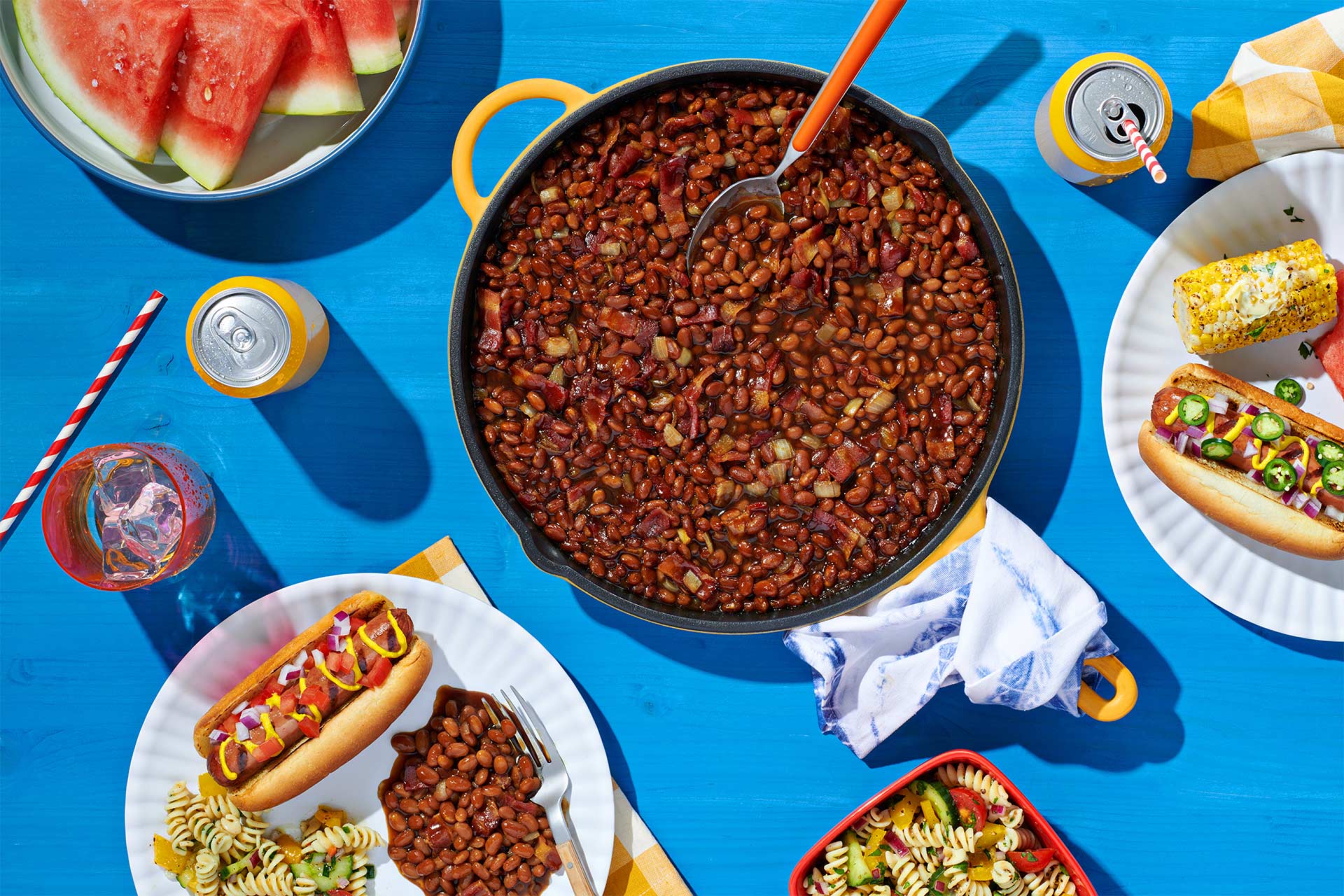 Top down view of baked beans (with bacon and onions) in an enamled pot surrouned by paper plates with hot dogs, pasta salad, corn on the cob elote, and watermelon. 