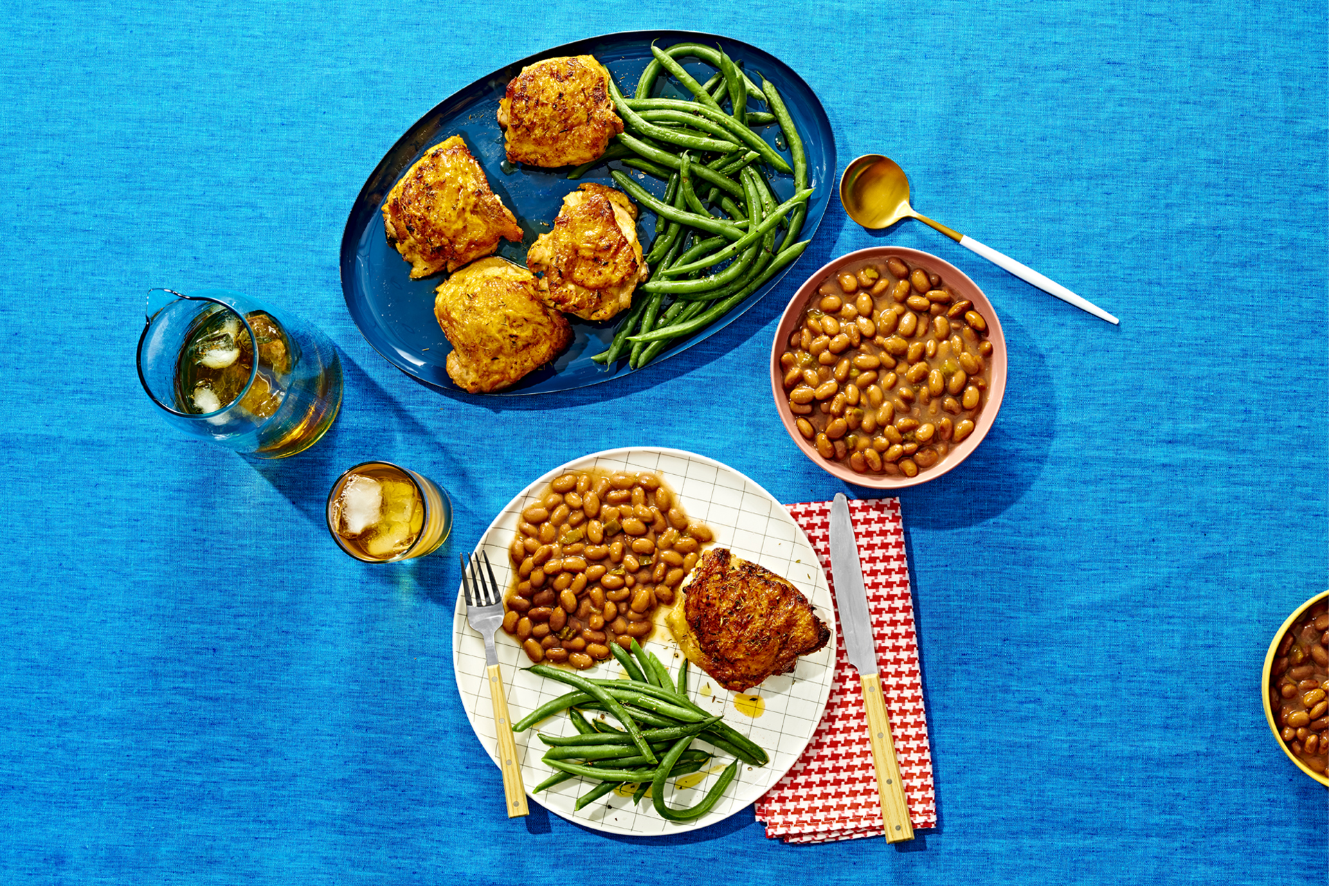 A dinner setup with a loaded plate dinner plate, a serving plate with chicken and green beans, and bowl of pinto beans on a blue table cloth.