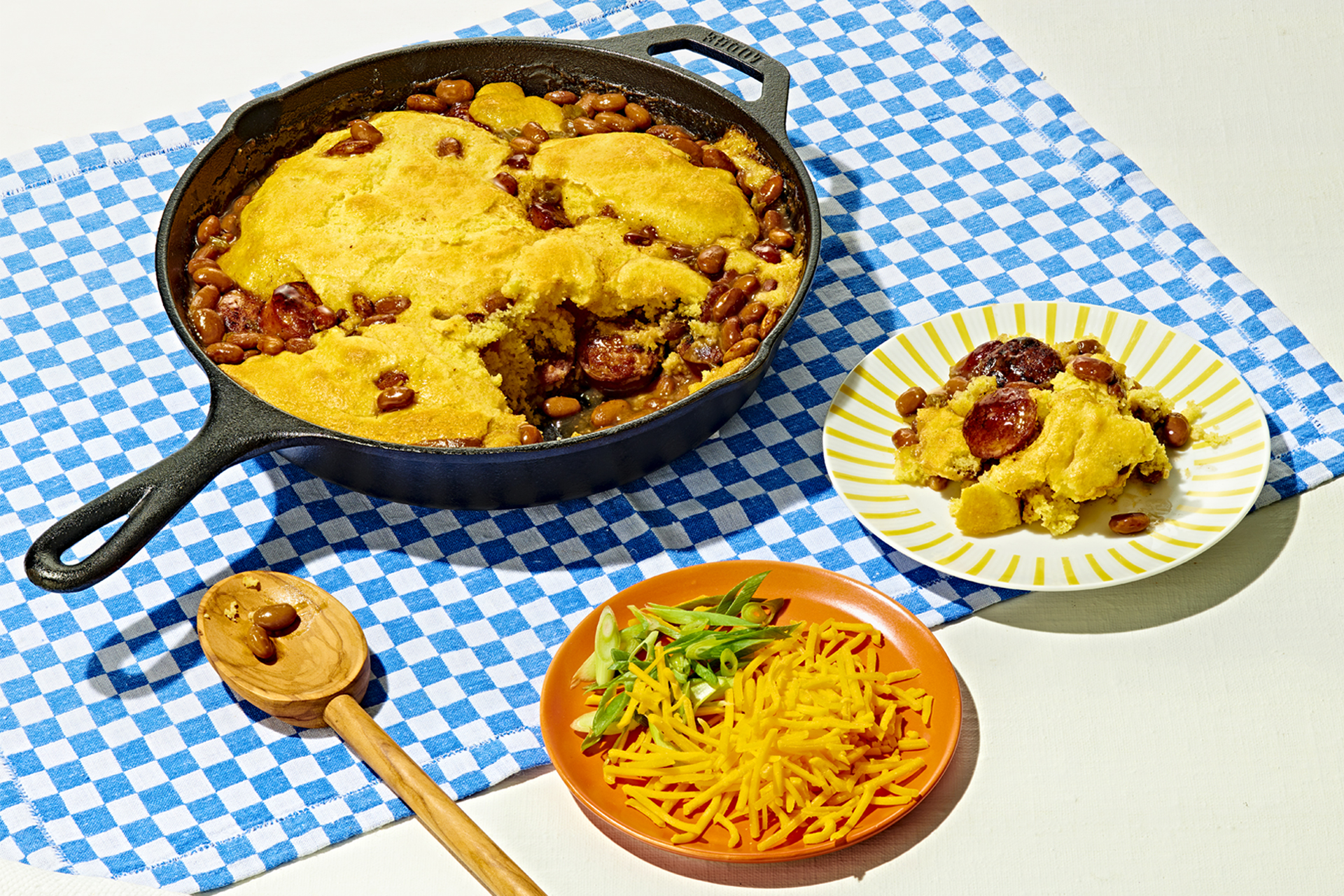 A cast iron skillet filled with a cornbread bake next to loaded plates on a blue gingham table cloth.