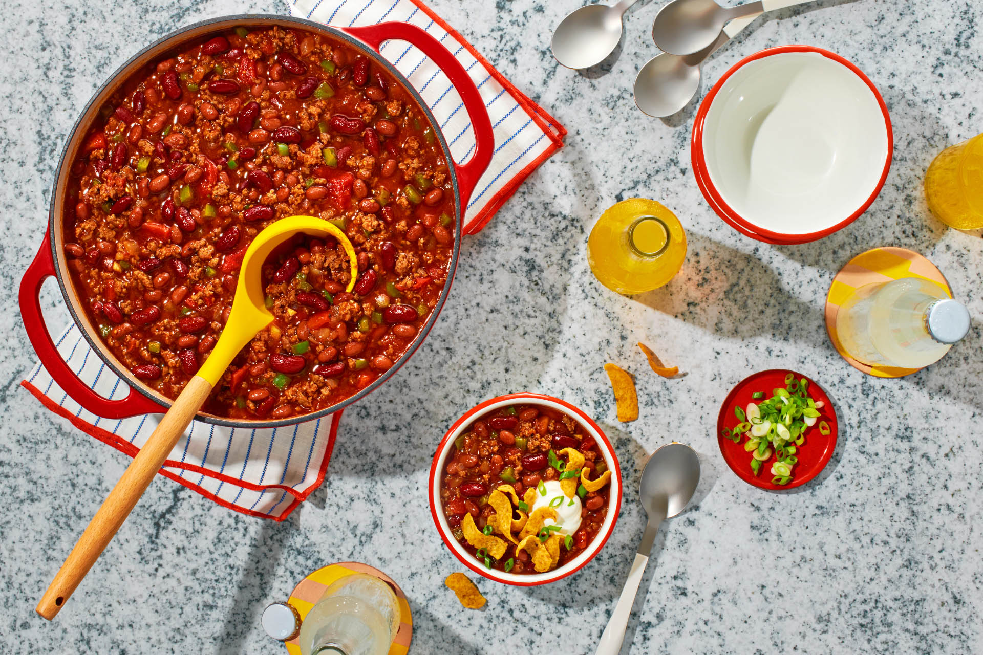Overhead view of a big pot of chili on a table alongside a bowl of chili, drinks and silverware.