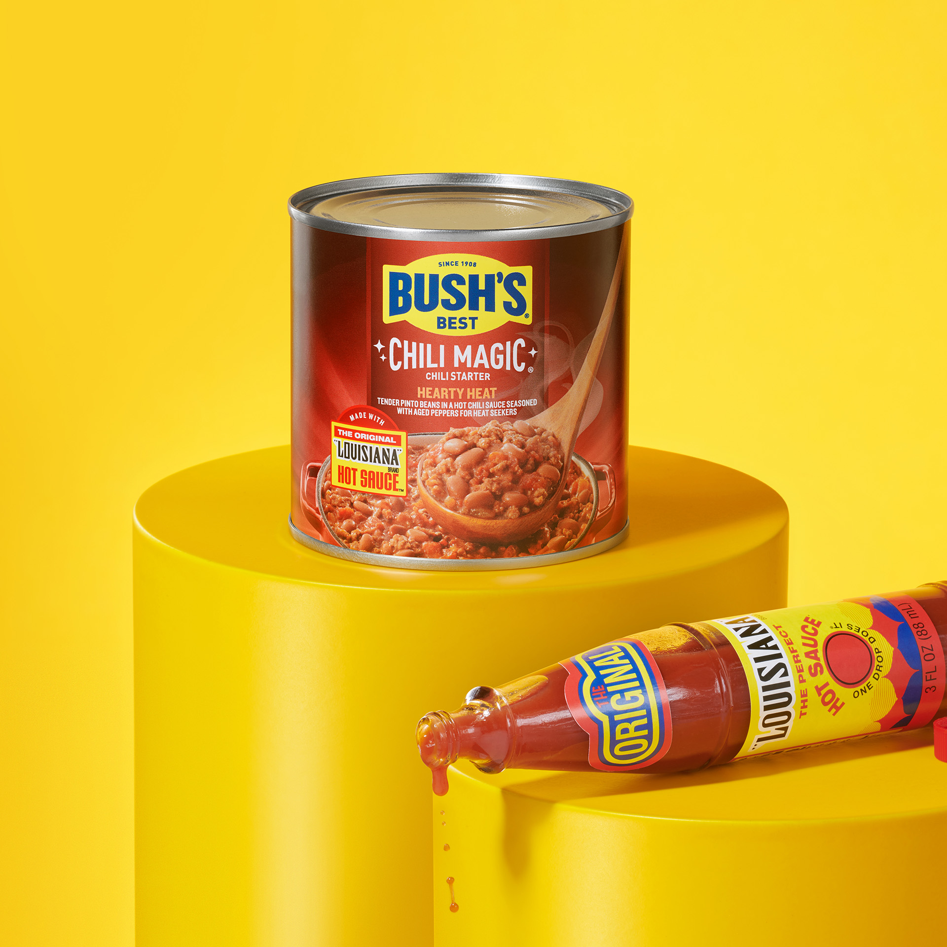 Can of Bush’s Chili Magic Hearty Heat on a pedestal with a bottle of hot sauce.