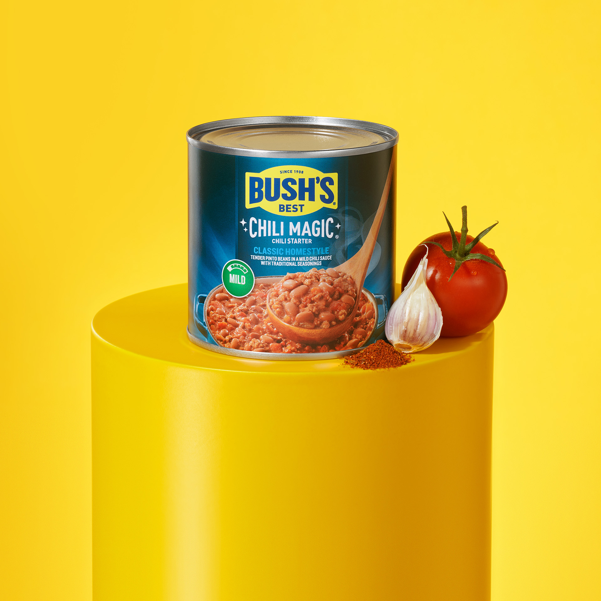 Can of Bush’s Chili Magic Classic Homestyle on a pedestal with a tomato, garlic clove and spices.