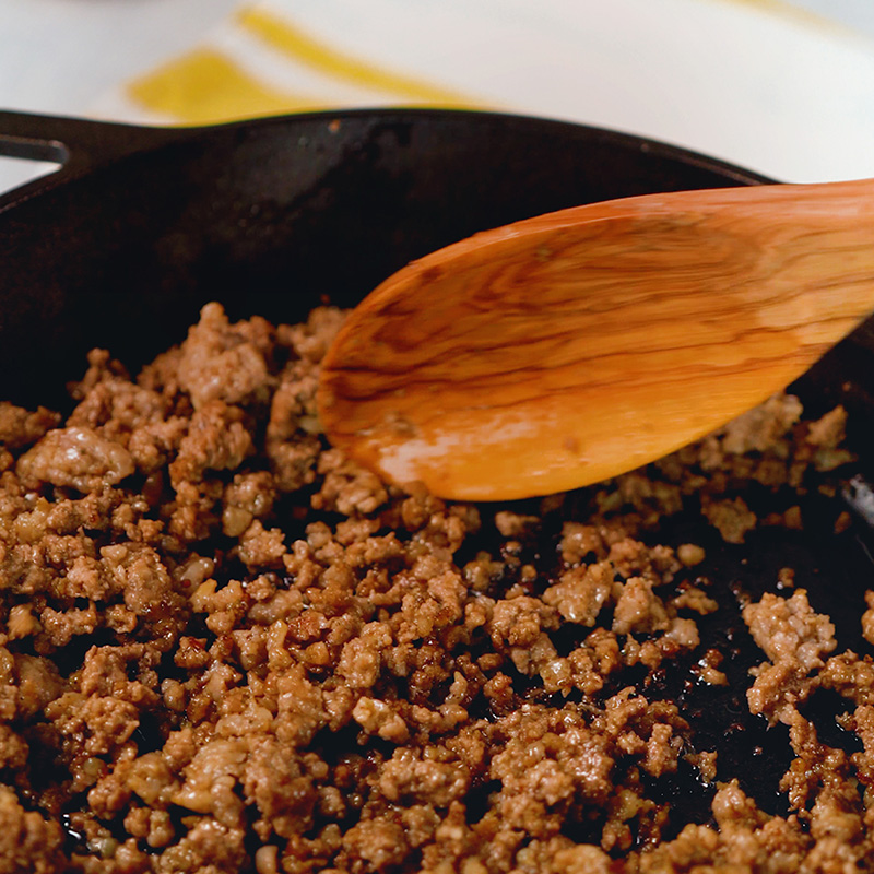 Cast iron pan full of ground beef being stirred with a wooden spoon.