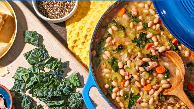 Hearty white bean and kale soup in a blue pot with fresh ingredients on the side.