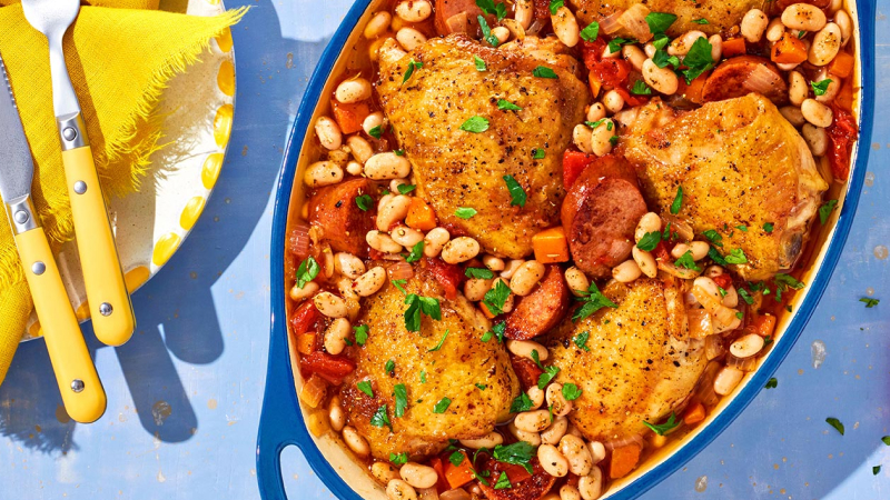 Blue baking dish filled with roasted chicken thighs, white beans, sausage, and vegetables, garnished with fresh parsley.