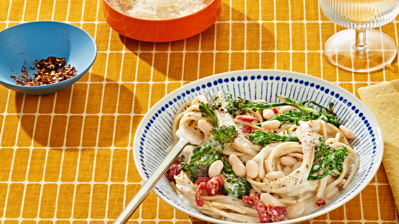 White bean and vegetable pasta in a creamy sauce served in a blue-striped bowl on a yellow patterned tablecloth, with a sprinkle of chili flakes on the side.