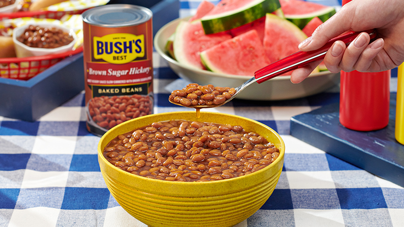 A spoonful of baked beans being held above a yellow bowl filled with beans, with a can of Bush's<sup>®</sup> Brown Sugar Hickory Baked Beans and sliced watermelon in the background on a checkered tablecloth.