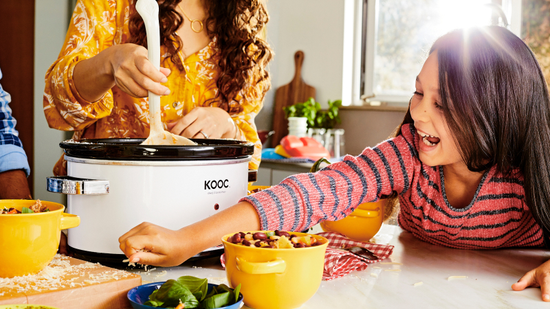 Smiling girl reaches across counter to grab cheese while her mom stirs chili in an instant pot.