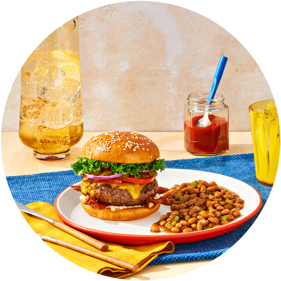 A plate with a burger and baked beans with a can of Bush's Zero Sugar Added Baked Beans on a picnic table.
