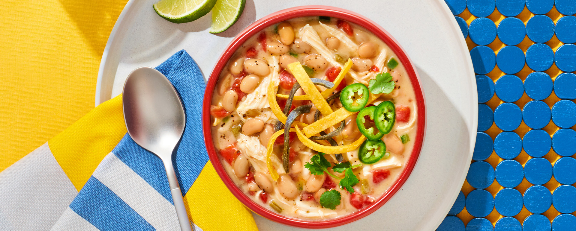 White chicken chili topped with jalapenos and tortilla strips in a red bowl on a white plate with spoon, lime slices and a striped napkin.