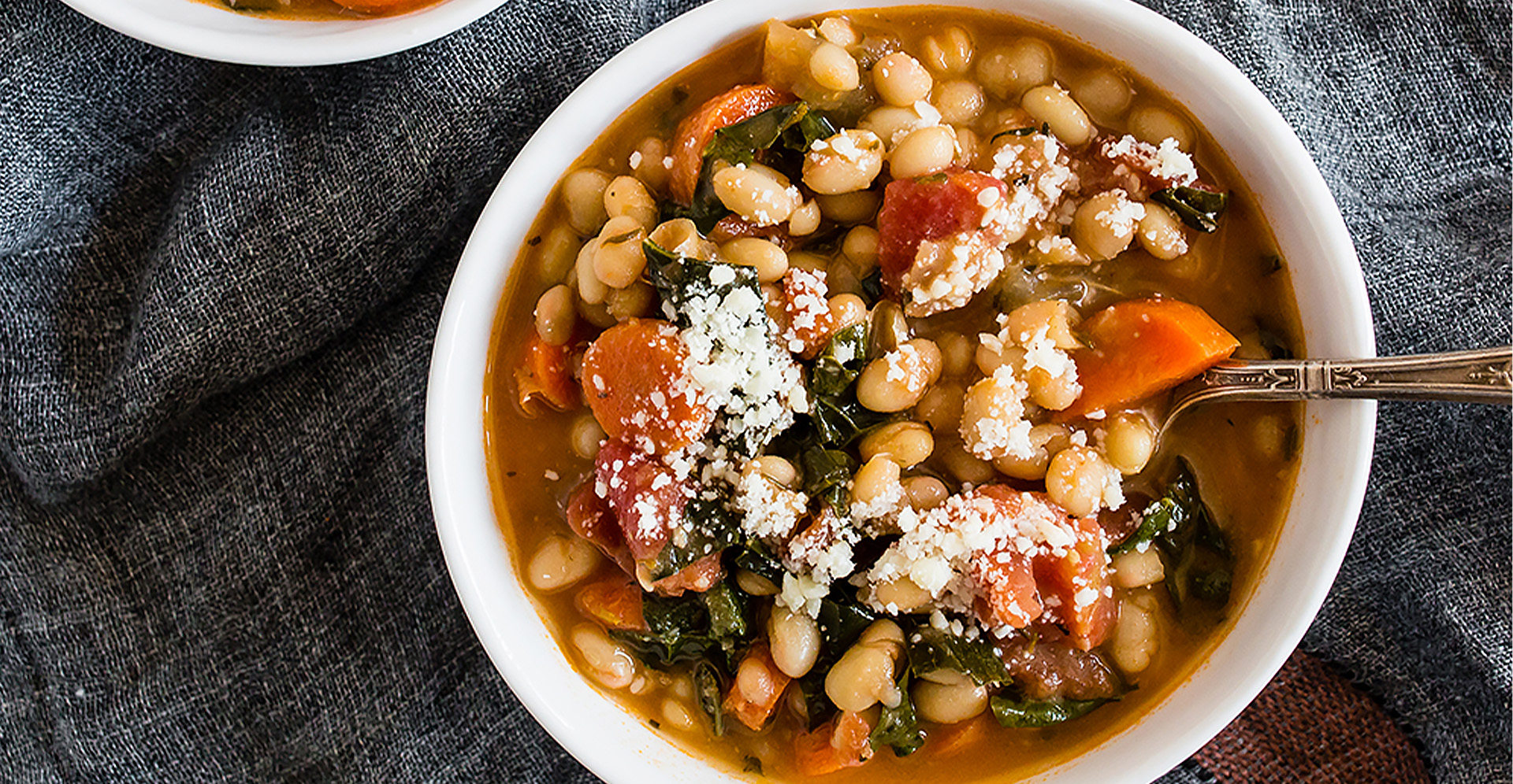 Tuscan tomato and white bean soup with kale with spoon in white bowl, garnished with parmesean cheese