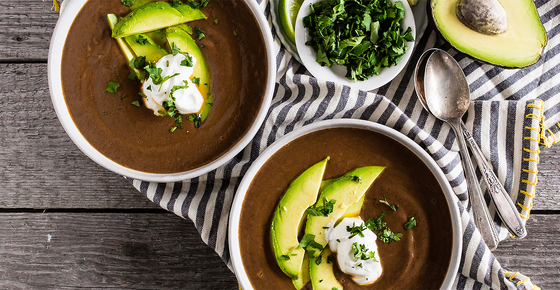 Creamy black bean soup in two white bowls with sour cream and avocado on top