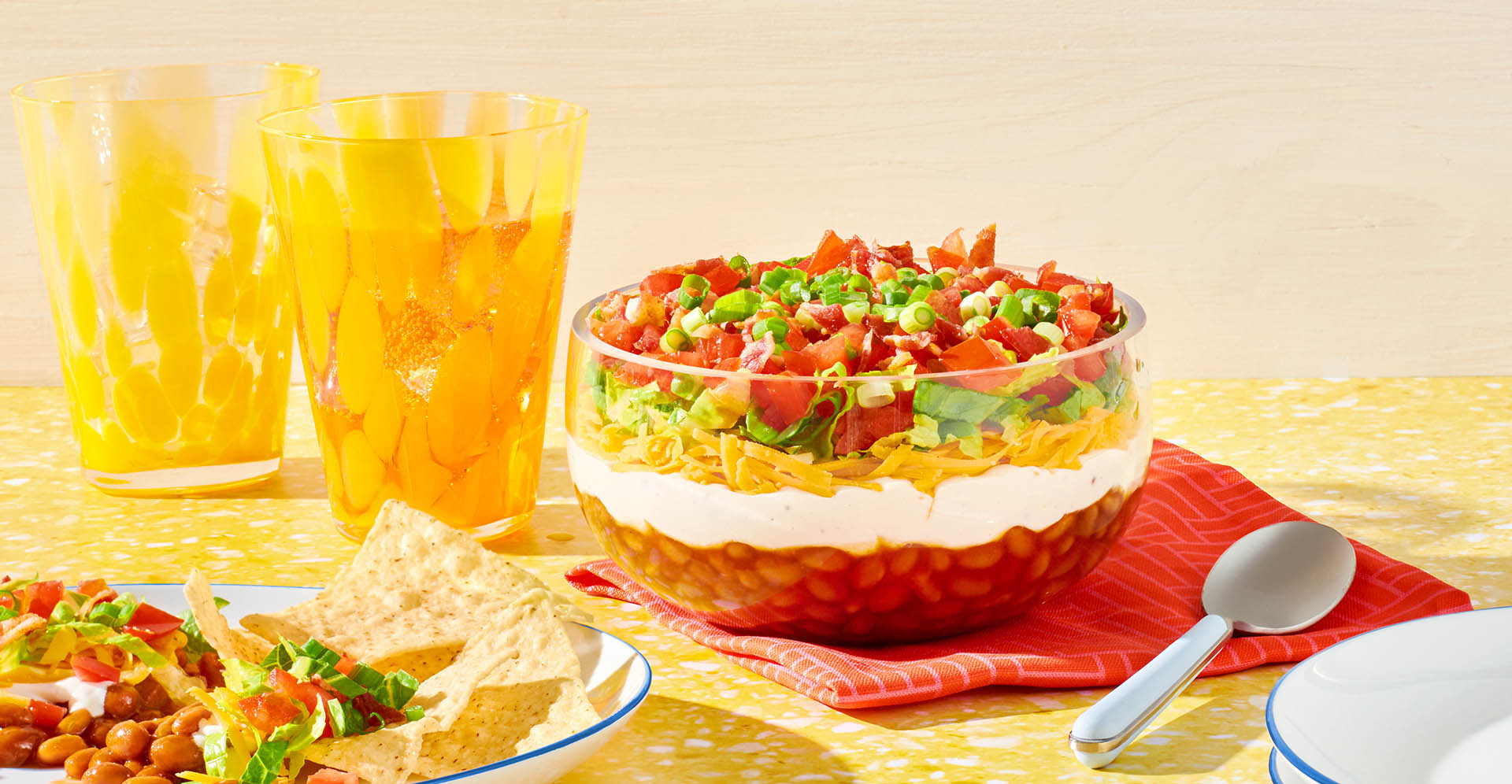 A bowl of layered bean dip on a red napkin next cups.