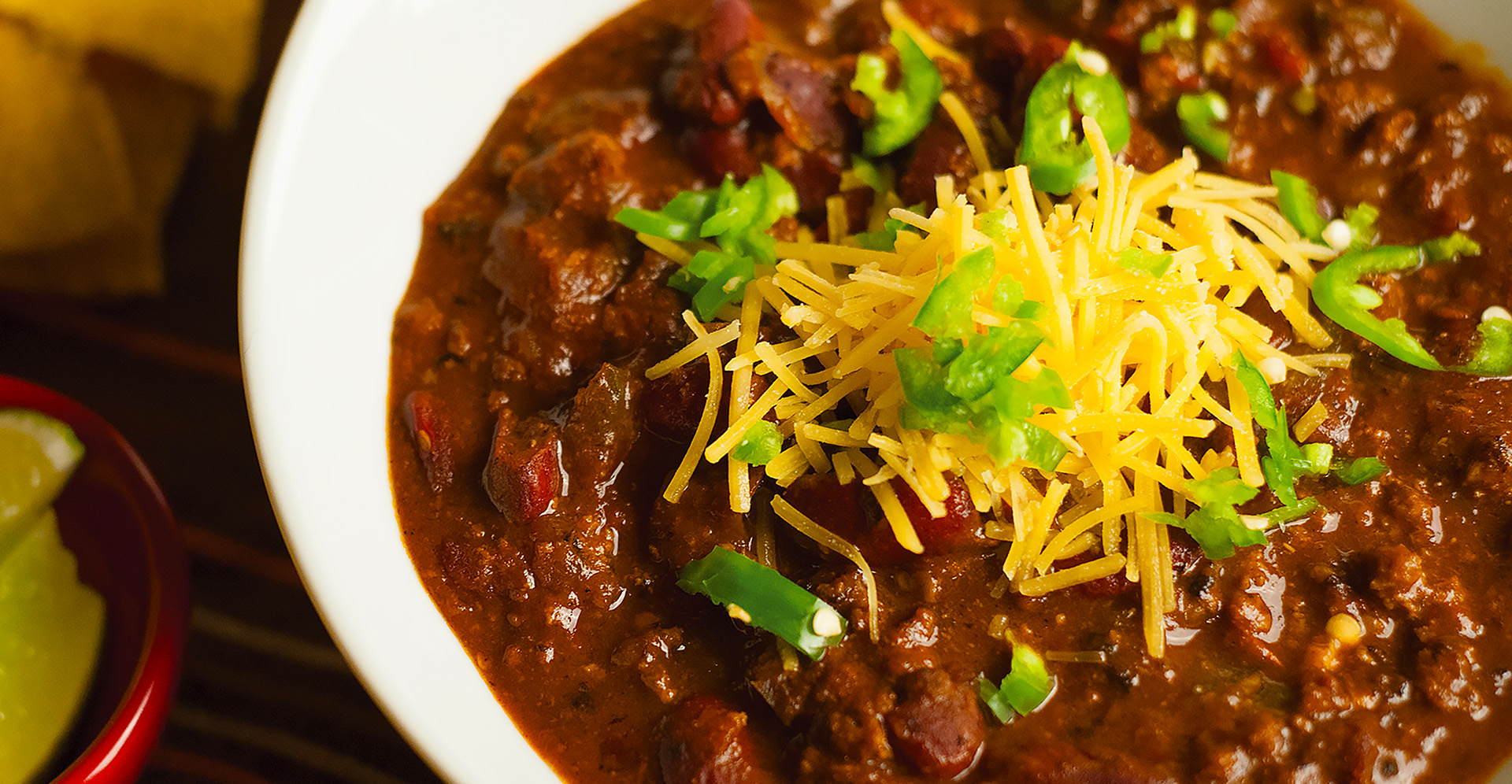 Close-up of beef and dark red kidney bean chili in white bowl, garnished with shredded cheddar cheese