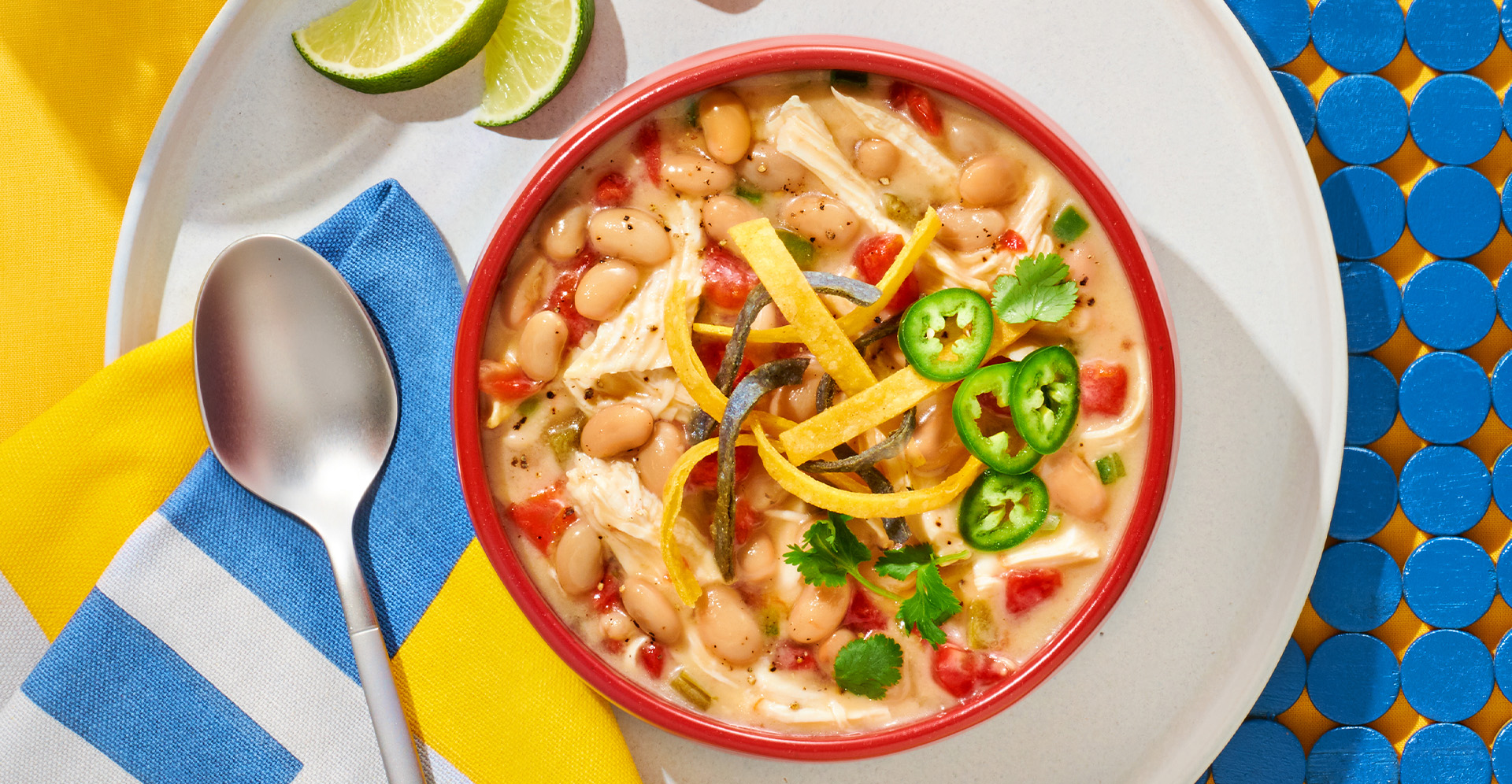 White chicken chili topped with jalapenos and tortilla strips in a red bowl on a white plate with spoon, lime slices and a striped napkin