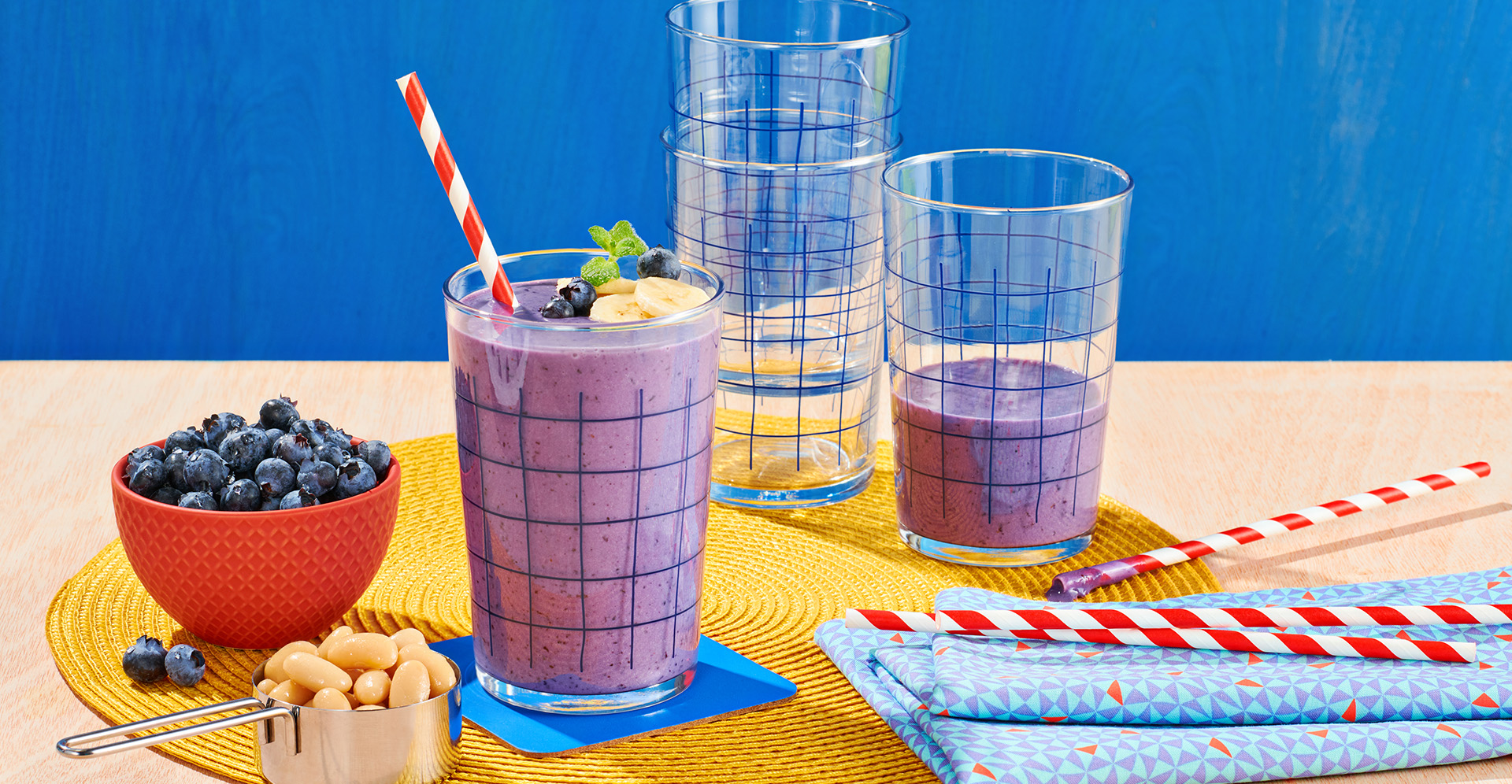 Banana blueberry and bean smoothie with striped straw next to a red bowl filled with blueberries and a silver measuring cup filled with white beans
