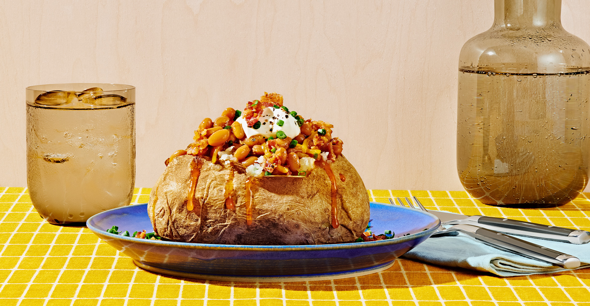 Baked potato topped with baked beans, sour cream and bacon bits on blue plate next to a glass of ice water and a fork and knife