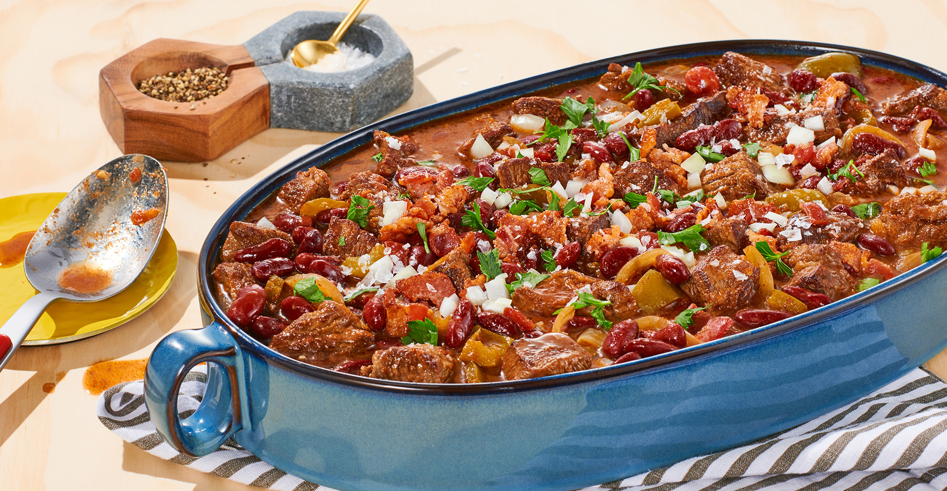 Braised beef and bacon chili garnished with onions in an oval blue pot