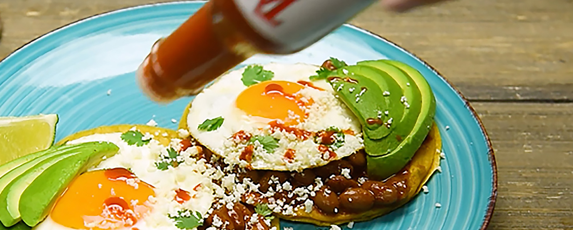 Someone pouring Tapitio hot sauce on breakfast tacos with beans, sunny side up eggs, avocado, tortillas, and lime slices