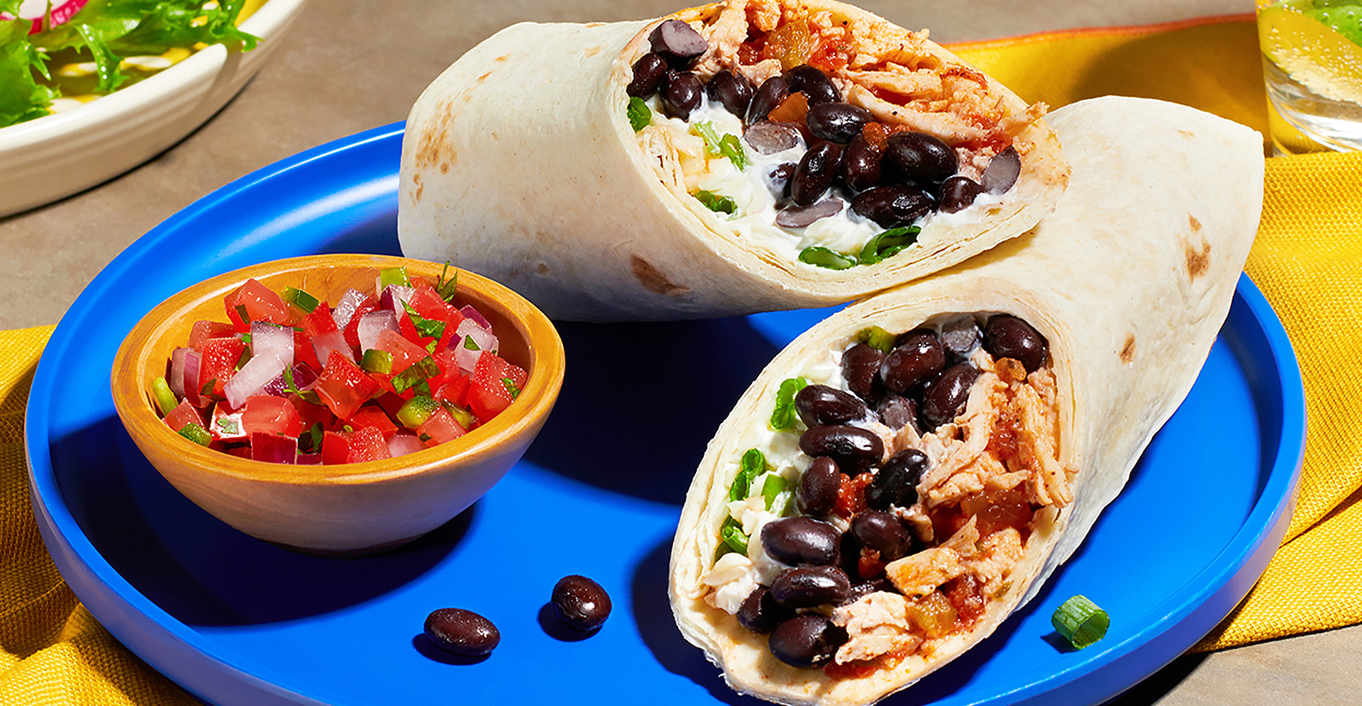 A chicken and black bean burrito sliced in half on a blue plate with small bowl of tomato salsa