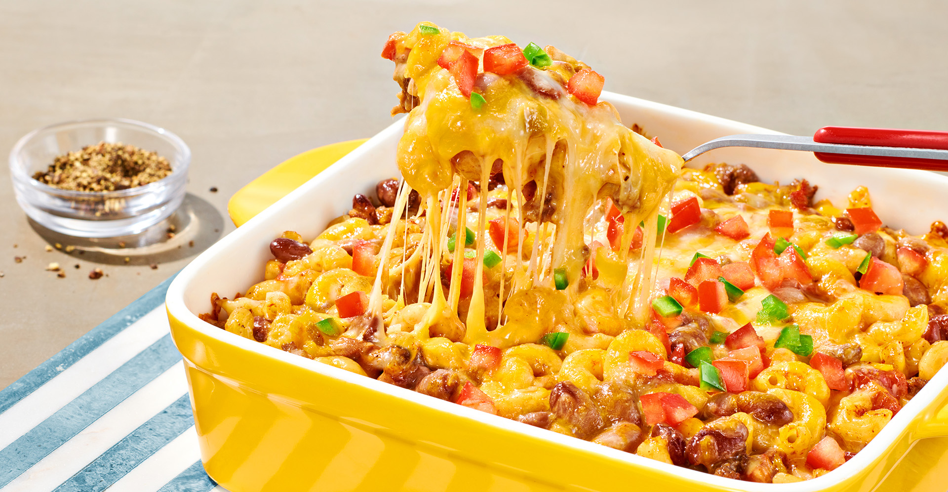 A spoon taking a scoop of a baked Mexican chili bean, macaroni and cheese casserole topped with tomato in yellow baking dish