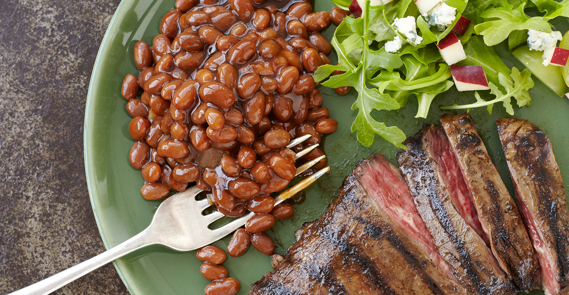 Sliced marinated steak with baked beans and a green salad with pear and bleu cheese on a white plate