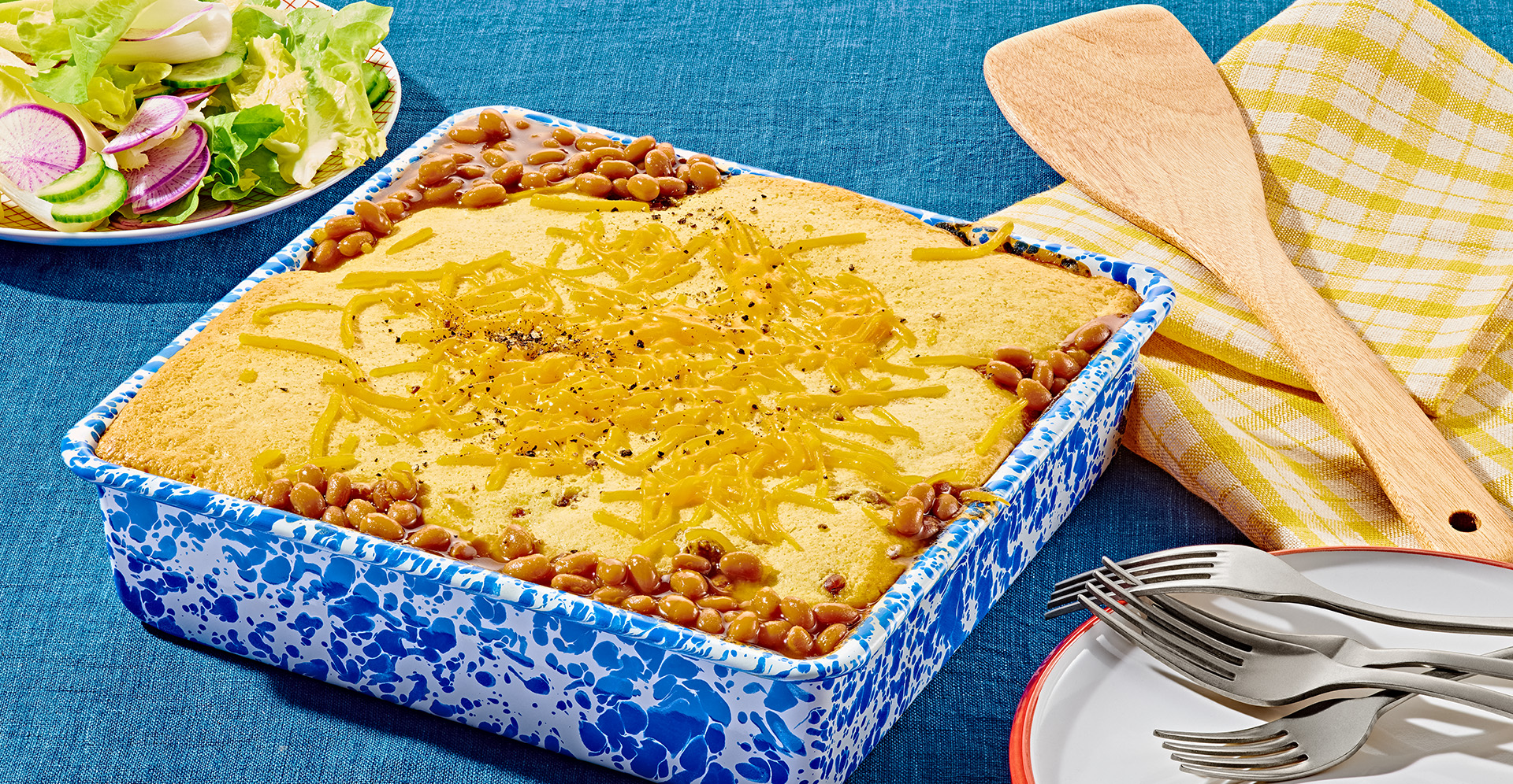 Baked bean, beef and cornbread casserole topped with melted cheese in square blue and white patterned baking dish