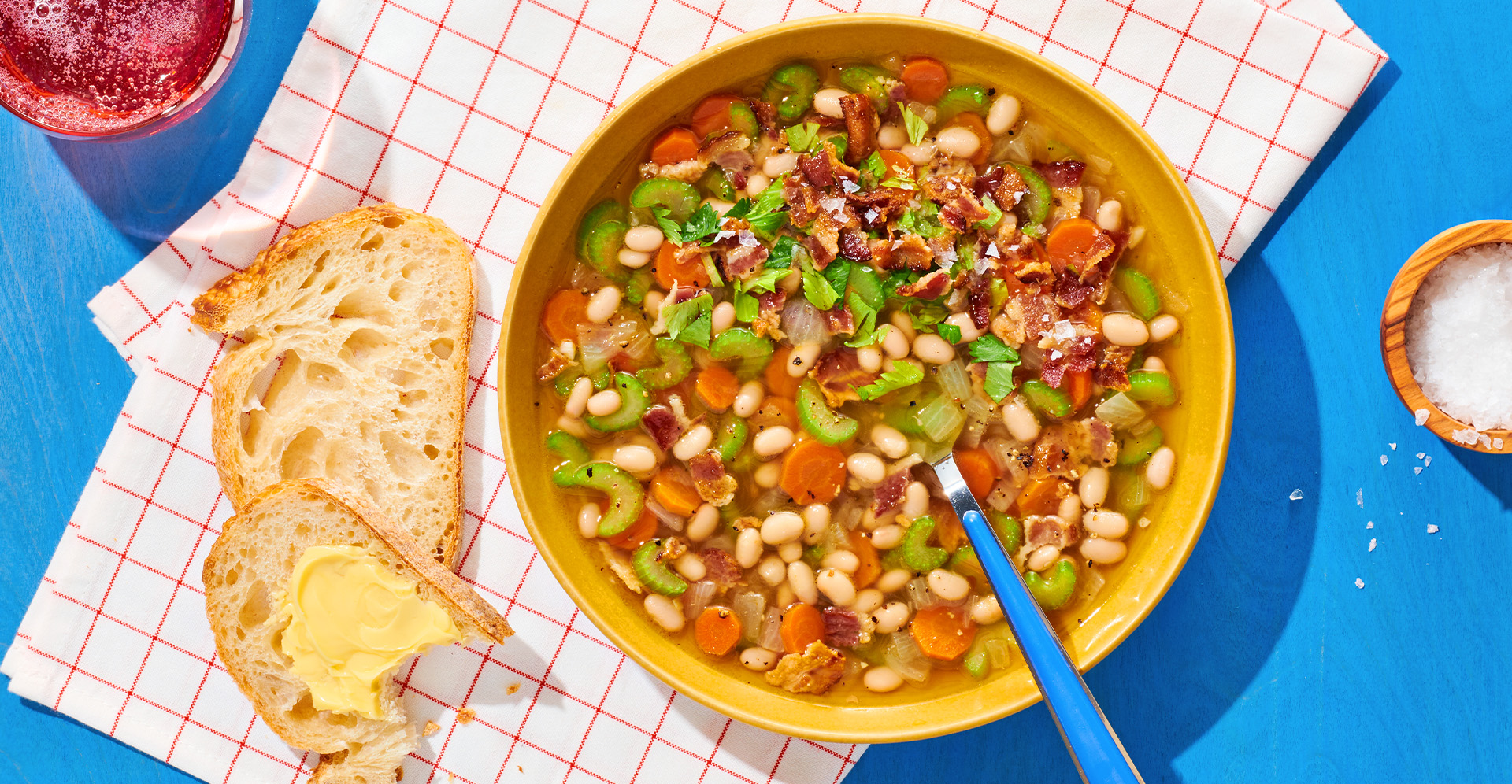 Bowl of white bean soup with bacon, carrot, celery in a yellow bowl. Buttered bread and salt on the side.
