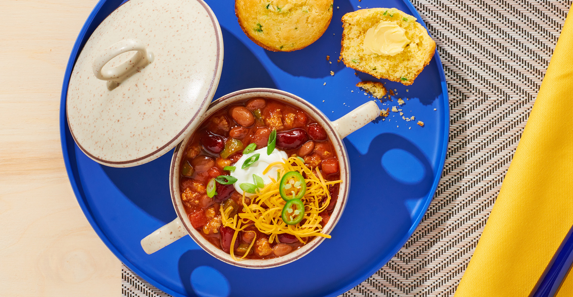 A bowl of chili topped with sour cream, jalapenos, and shredded cheese on a blue tray with corn muffins