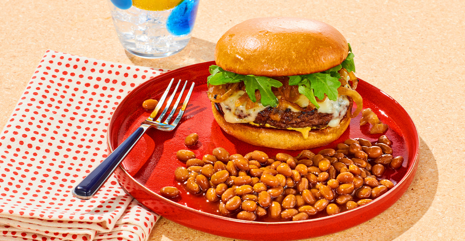 A French onion burger topped with cheese and greens with a side of baked beans on a red plate