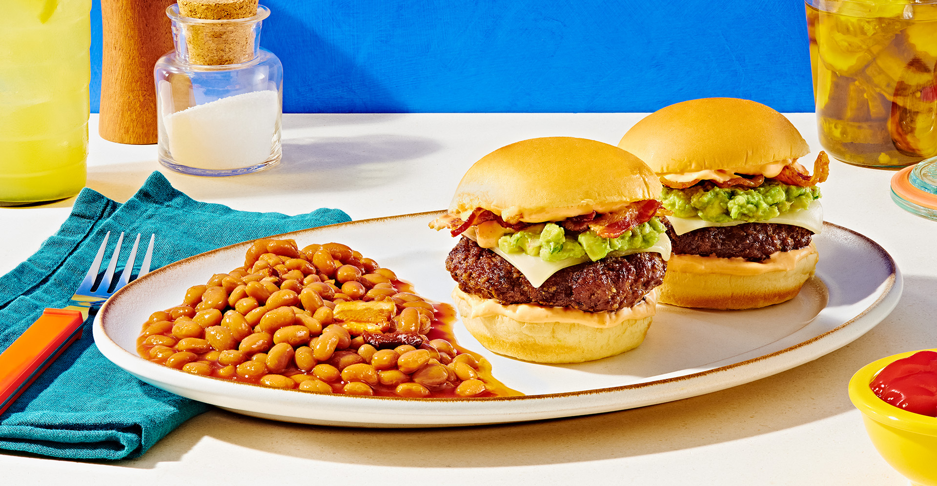 Two hamburgers topped with guacamole and mozerella with a side of baked beans with bacon on white platter