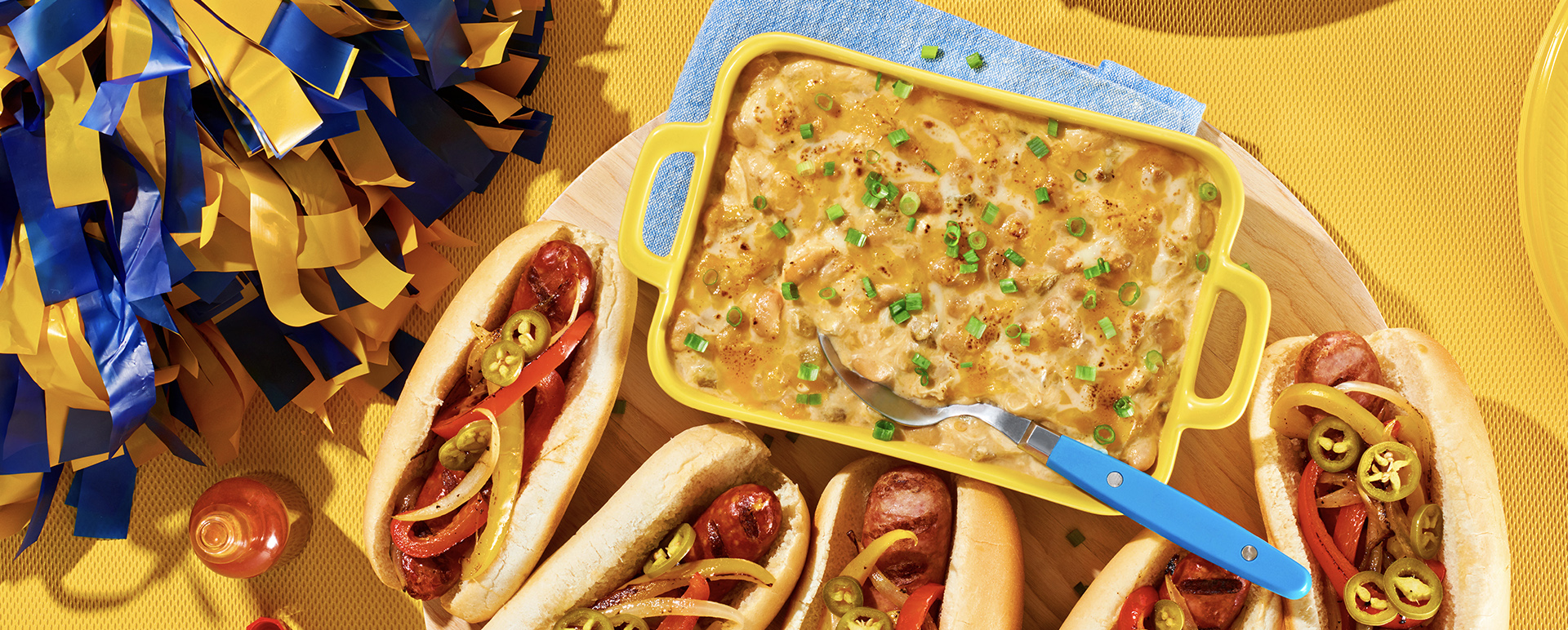 White chicken chili dip in a yellow casserole next to hotdogs and near a bowl of carrot sticks.