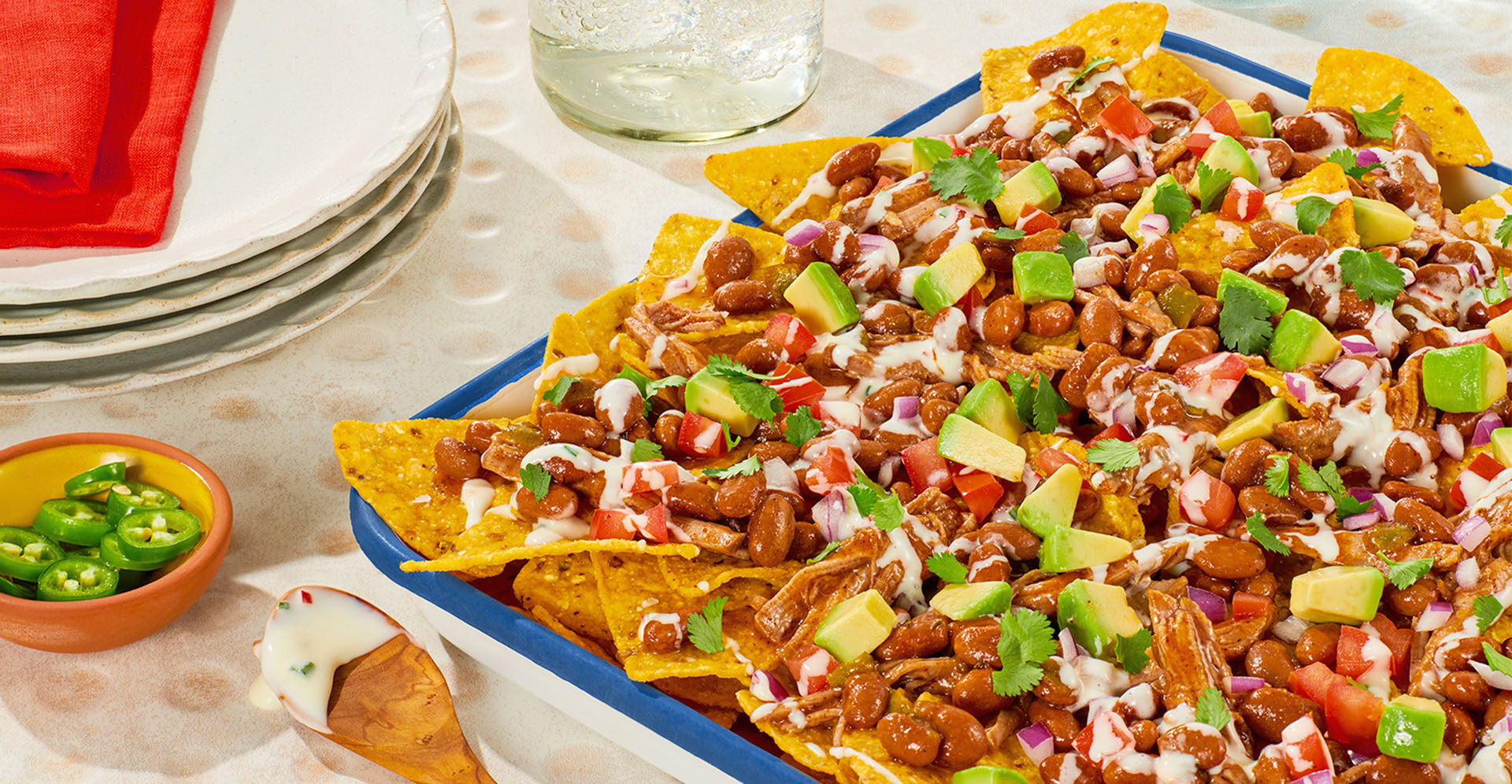 A casserole dish full of nachos siting on a table next to several plates and a drink.