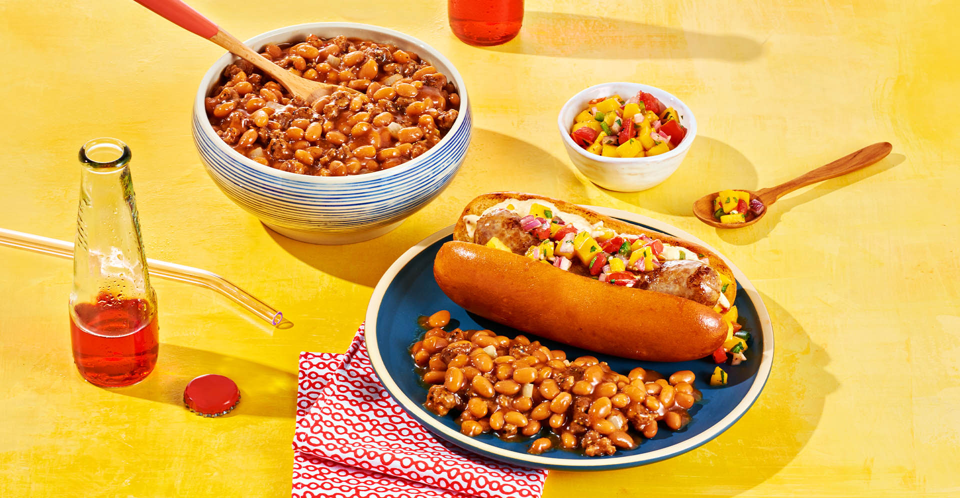 A grilled brat on a bun with a side of hamburger baked beans next to bowl of hamburger baked beans.