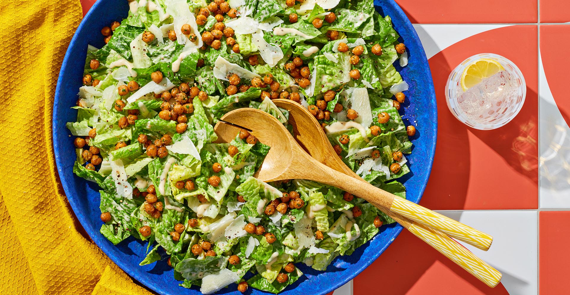 Large blue bowl filled with grilled caesar salad atop a red and white tile table with a yellow napkin.