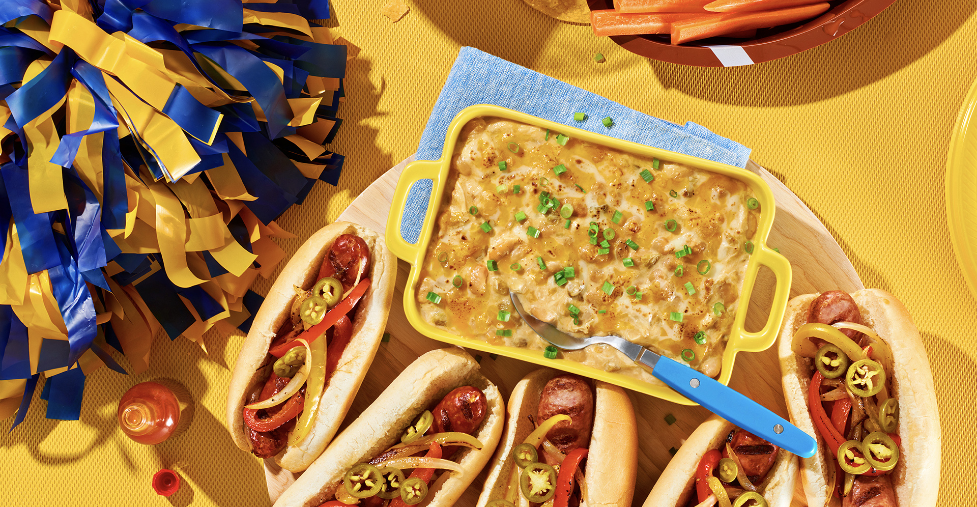 White chicken chili dip in a yellow casserole next to hotdogs and near a bowl of carrot sticks. 